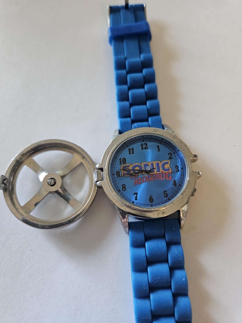 Puede incluir: Un reloj azul de Sonic the Hedgehog con una correa azul a juego. La esfera del reloj presenta el logotipo de Sonic y un fondo azul. El reloj tiene una caja de metal plateado y una tapa con bisagras.