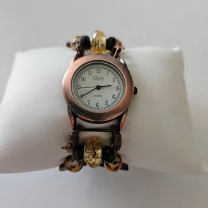 May include: A copper-toned wristwatch with a white face and black hands. The watch has a black and gold beaded band.