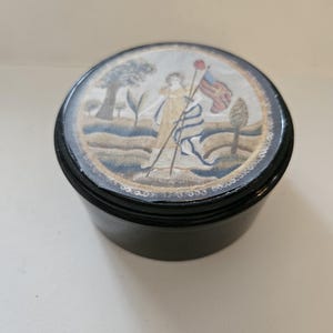 May include: A round, black container with a decorative lid. The lid features a painted scene of a woman holding a flag, with trees and a landscape in the background. The container is likely for storing small items.
