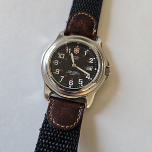 May include: A Swiss Army quartz wristwatch with a round silver-tone metal case and a black dial featuring white Arabic numerals, a date window, and luminous hands. The watch is attached to a black woven strap with brown leather accents.