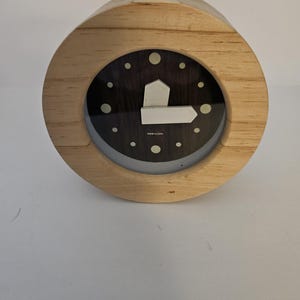 May include: A round wooden clock with a dark face and white hour markers. The clock has a light wood frame and a white arrow-shaped hand. The clock is a decorative item.