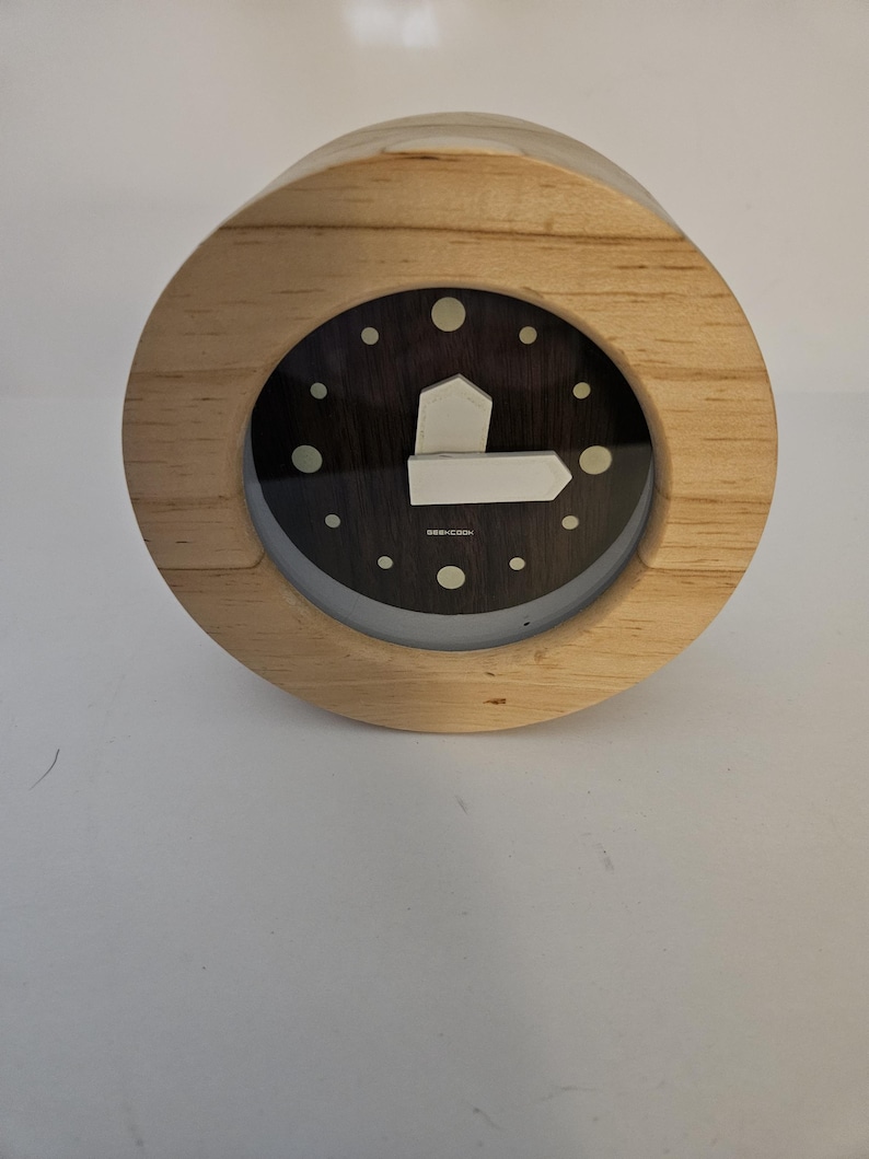 Puede incluir: Un reloj redondo de madera con una esfera oscura y marcadores de hora blancos. El reloj tiene un marco de madera clara y una manecilla blanca en forma de flecha. Es un objeto decorativo.
