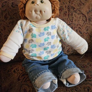 May include: A vintage doll with a soft, vinyl face, green eyes, and a tuft of brown yarn hair. The doll wears a white long-sleeved shirt with a pattern of blue and yellow elephants, and blue denim jeans.