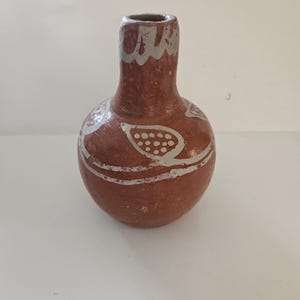 May include: A small, round, terracotta-colored vase with a narrow neck and a silver rim. The vase is adorned with white, hand-painted, abstract designs. The overall aesthetic is rustic and handcrafted, suitable for displaying small flowers or as a decorative piece.