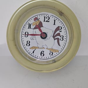 May include: A round wall clock with a cartoon rooster design. The clock face is white with black numbers and a red second hand. The clock has a light yellow frame.