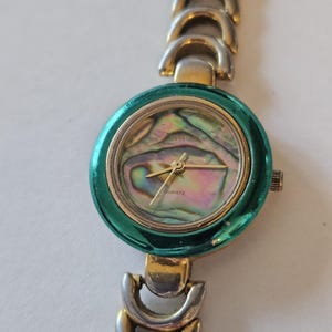 May include: A wristwatch featuring a round, metallic teal bezel and an iridescent abalone shell dial. The watch has gold-tone hands, the word QUARTZ printed on the face, and a two-tone silver and gold link bracelet with curved, crescent-shaped segments.