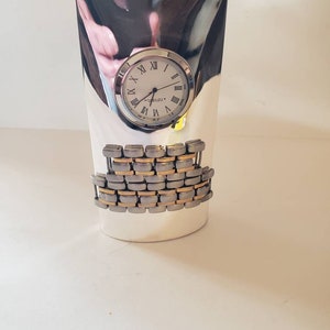 May include: Silver and gold tone clock with Roman numeral hour markers and a silver band around the face. The clock is on a silver stand with a silver and gold tone band around the middle.