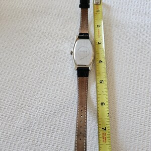 May include: A silver rectangular watch with a brown leather strap. The watch face is not visible. The back of the watch has the number "23238" printed on it. The watch is measured with a yellow tape measure.