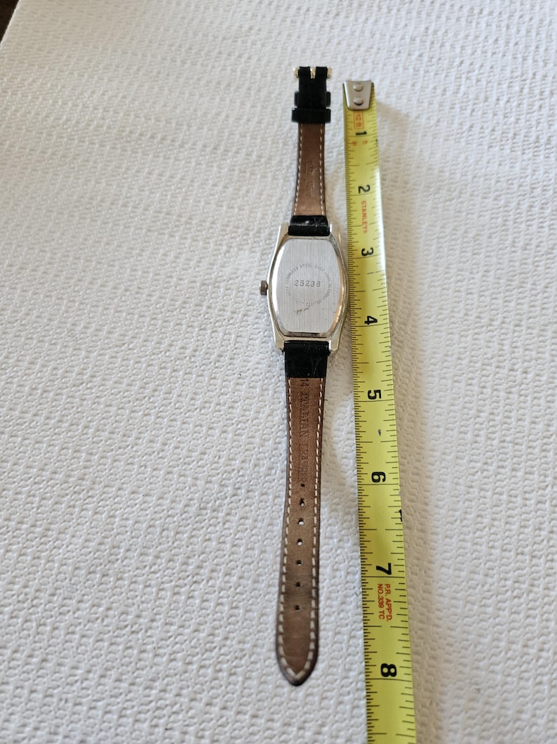 May include: A silver rectangular watch with a brown leather strap. The watch face is not visible. The back of the watch has the number "23238" printed on it. The watch is measured with a yellow tape measure.