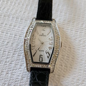 May include: A rectangular silver quartz watch with a white face and crystal-set bezel. The watch has black Roman numerals and hands, with a black leather strap. The brand name "minicci" is visible on the watch face.