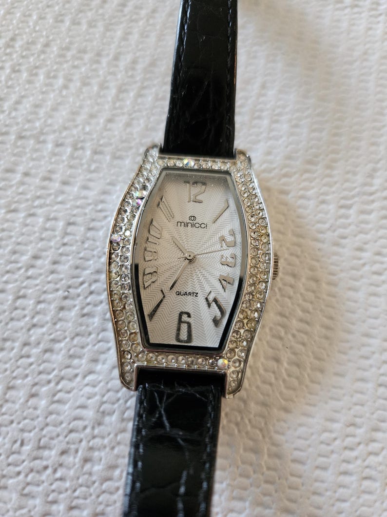 May include: A rectangular silver quartz watch with a white face and crystal-set bezel. The watch has black Roman numerals and hands, with a black leather strap. The brand name "minicci" is visible on the watch face.