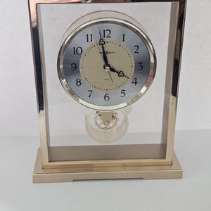 May include: A gold-toned rectangular mantel clock with a clear glass frame. The clock face is silver with black numerals and hands. The clock has a pendulum and the brand name "HOWARD MILLER" is visible.