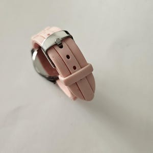 Op de afbeelding: Een close-up van een roze polshorloge met een zilveren gesp. Het horloge heeft een lichtroze siliconen band. De wijzerplaat is zichtbaar, met een zilveren rand. Het horloge staat tegen een witte achtergrond.