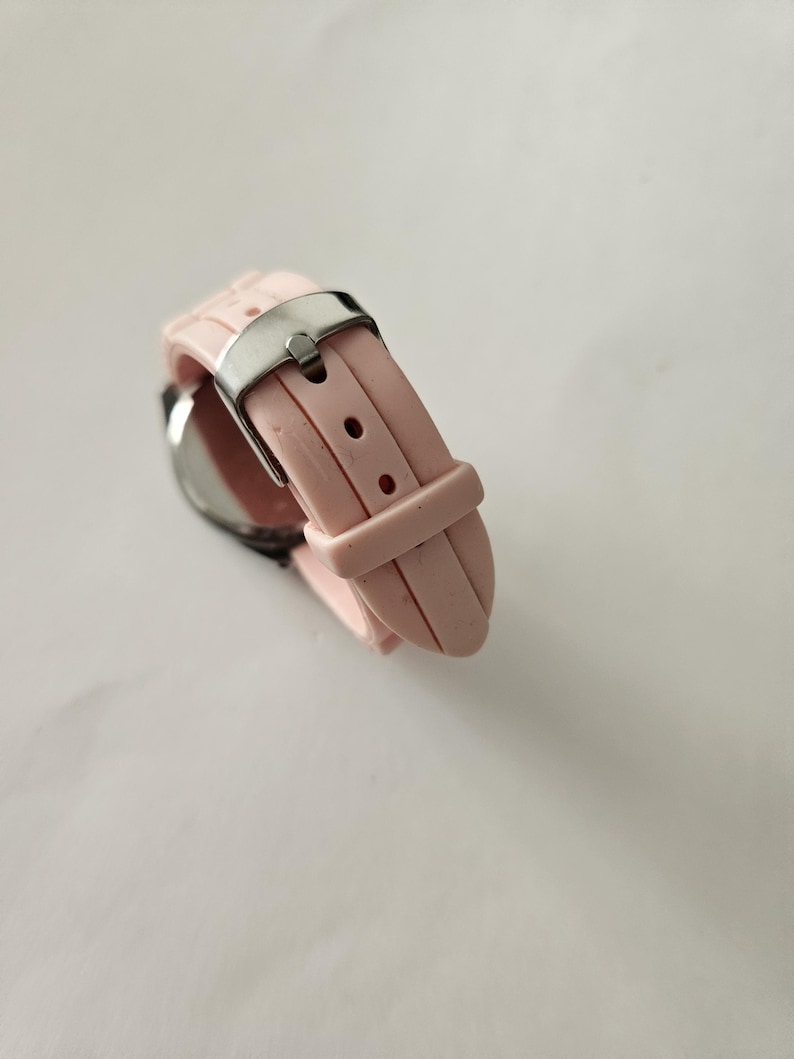 Op de afbeelding: Een close-up van een roze polshorloge met een zilveren gesp. Het horloge heeft een lichtroze siliconen band. De wijzerplaat is zichtbaar, met een zilveren rand. Het horloge staat tegen een witte achtergrond.