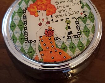 May include: A round, silver pill box with a colorful design featuring a woman with orange hair and a red dress. The design includes the text "Some days should come with a delete button!"