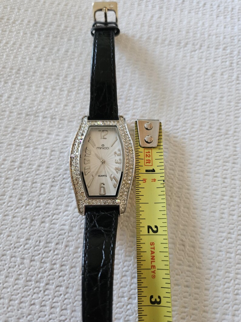 May include: A rectangular quartz watch with a silver bezel set with rhinestones. The watch face is white with black numerals and hands. The black leather strap is approximately 20 cm long.