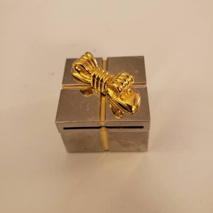 May include: A small, silver-colored box with a gold-colored bow on top. The box is square-shaped and has a metallic finish. The bow is intricately detailed, adding a touch of elegance to the overall design. The box appears to be a decorative item or a small gift container.