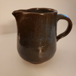 May include: A small, brown ceramic pitcher with a slightly uneven rim and a handle. The pitcher has a speckled glaze with a blue-grey hue.