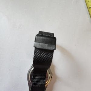 May include: A black wristwatch with a fine mesh metal strap and a rectangular clasp, positioned next to a yellow measuring tape showing markings in inches. The watch case is silver-toned on the back, and the strap features a textured pattern.