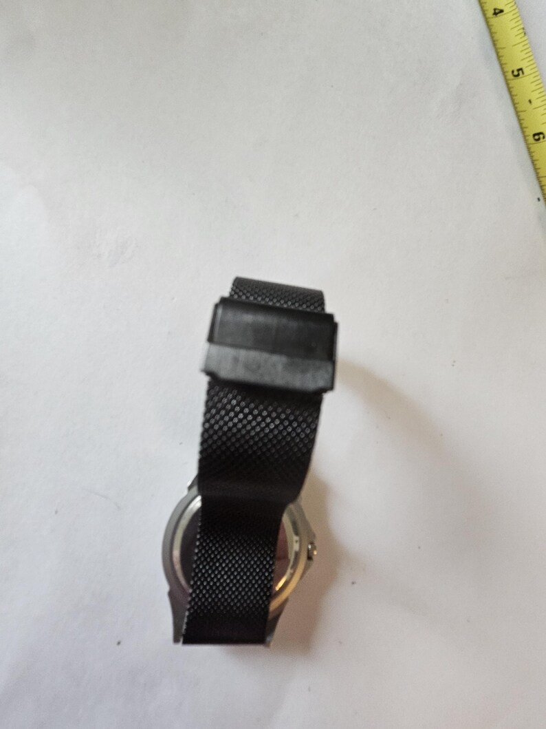 May include: A black wristwatch with a fine mesh metal strap and a rectangular clasp, positioned next to a yellow measuring tape showing markings in inches. The watch case is silver-toned on the back, and the strap features a textured pattern.