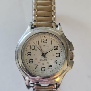 May include: A silver-toned quartz watch with a white face and black numerals. The watch has a stretch band and a round face with the word "QUARTZ" printed on the face. The watch hands are black.