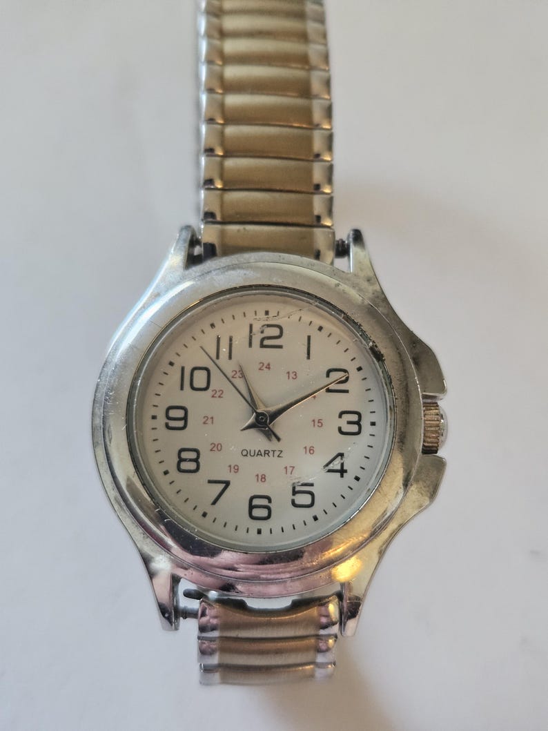 May include: A silver-toned quartz watch with a white face and black numerals. The watch has a stretch band and a round face with the word "QUARTZ" printed on the face. The watch hands are black.