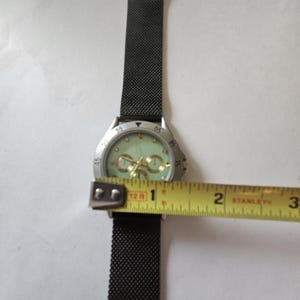 May include: A wristwatch with a black mesh strap and a silver-tone bezel featuring a light green dial with gold-tone accents. A yellow measuring tape is positioned across the watch face, showing a diameter of approximately 1.5 inches.