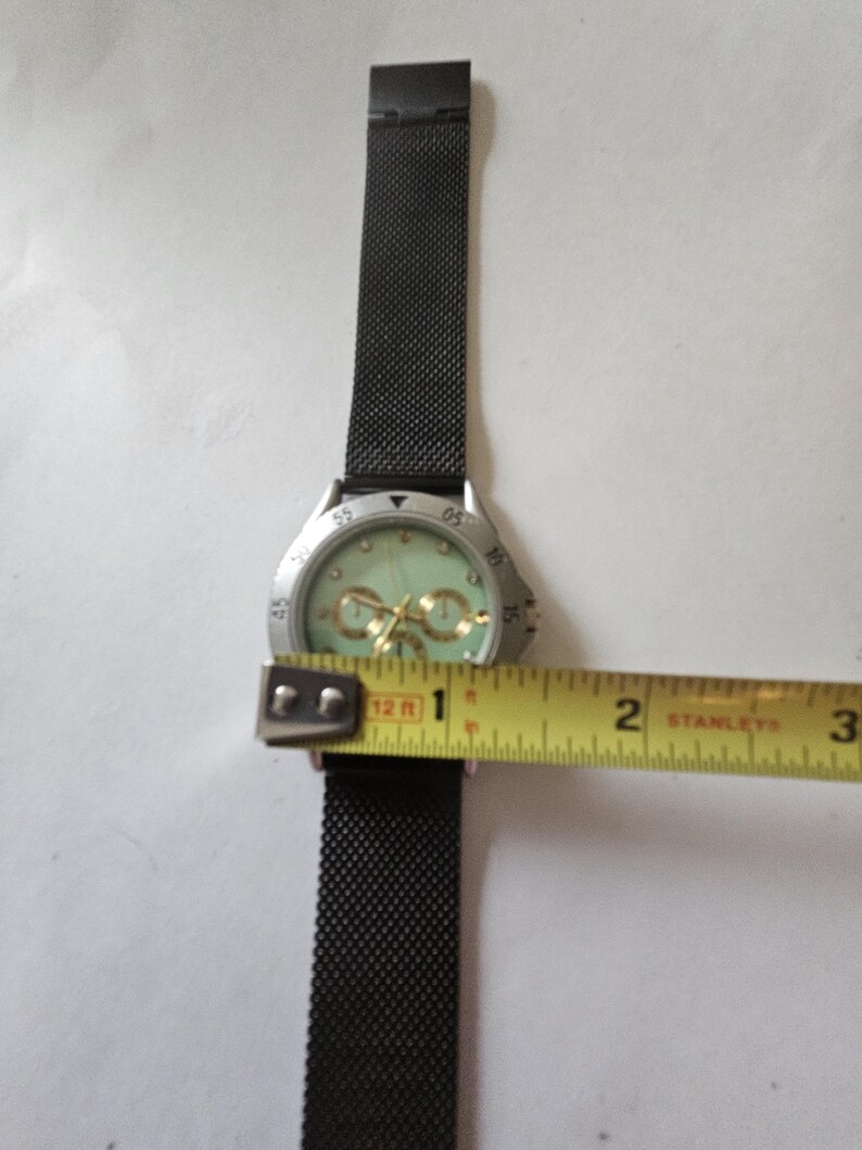 May include: A wristwatch with a black mesh strap and a silver-tone bezel featuring a light green dial with gold-tone accents. A yellow measuring tape is positioned across the watch face, showing a diameter of approximately 1.5 inches.