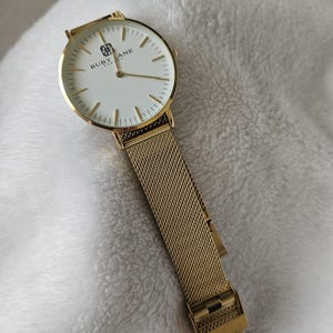 May include: A gold-tone watch with a white face and a mesh metal band. The watch face has the text "RUBY LANE" printed on it.