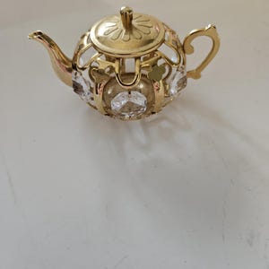 May include: A decorative gold-colored teapot with a filigree design and clear crystal accents. The teapot features a lid with a floral design and a curved handle. The spout is slender and elegant. The teapot is a decorative item.