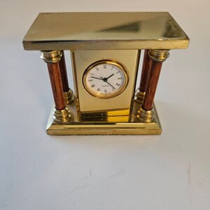 May include: A small gold-toned clock with a white face and black hands. The clock is on a brass stand with two wooden columns.