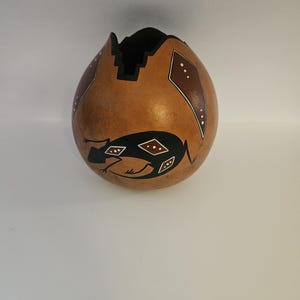 May include: A handcrafted gourd vase with a brown exterior and a black interior. The gourd features geometric designs in black, white, and brown, including a stylized frog. The top edge is cut in a decorative pattern.