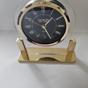 May include: A gold-toned Danbury Quartz clock with a black face and gold hands. The clock has Roman numerals and is set in a clear, arched frame. The base is rectangular and gold-toned.