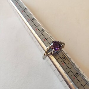 May include: A silver ring with a large, faceted, purple gemstone surrounded by smaller, clear gemstones. The ring is sized on a ring sizer that shows the size is 18.