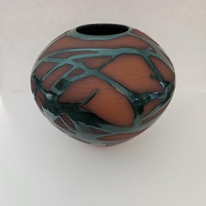 May include: A spherical ceramic vase with a matte terracotta-colored base and a glossy, dark teal glaze pattern that resembles interconnected branches or veins wrapping around the exterior surface, featuring a circular opening at the top.