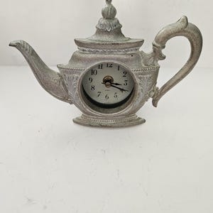 May include: A decorative teapot-shaped clock in a distressed silver finish. The clock face is visible within the teapot's body, displaying black numerals and hands. The teapot features a spout, handle, and lid, all with intricate detailing.