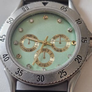 May include: A wristwatch with a mint green face, gold-tone hands, and crystal hour markers. The silver-tone bezel features black numerals from 05 to 55. Three gold-tone sub-dials display the day, date, and a 24-hour clock. A black mesh strap is attached.