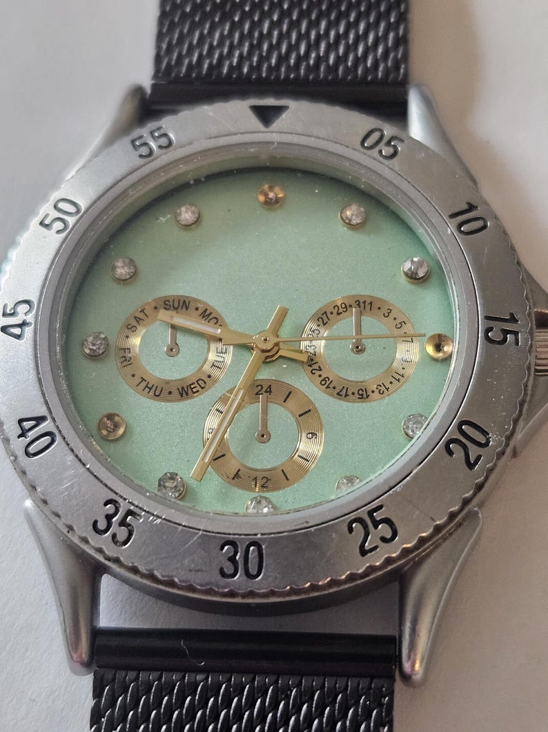 May include: A wristwatch with a mint green face, gold-tone hands, and crystal hour markers. The silver-tone bezel features black numerals from 05 to 55. Three gold-tone sub-dials display the day, date, and a 24-hour clock. A black mesh strap is attached.