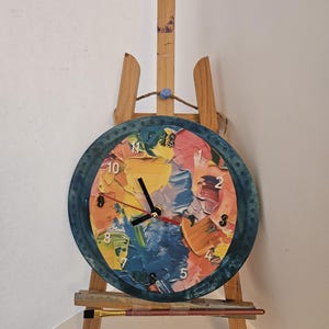 May include: A round wall clock with a colorful abstract painting design, set on a small wooden easel. The clock face features a textured, multi-colored pattern with black clock hands and numerals. A paintbrush rests on the easel's shelf.