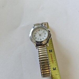 May include: A silver-toned wristwatch with a white face and black numerals. The watch has a stretch band and is measured with a yellow tape measure. The watch face reads "Quartz".