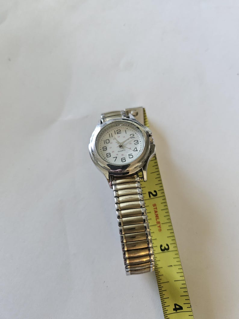 May include: A silver-toned wristwatch with a white face and black numerals. The watch has a stretch band and is measured with a yellow tape measure. The watch face reads "Quartz".