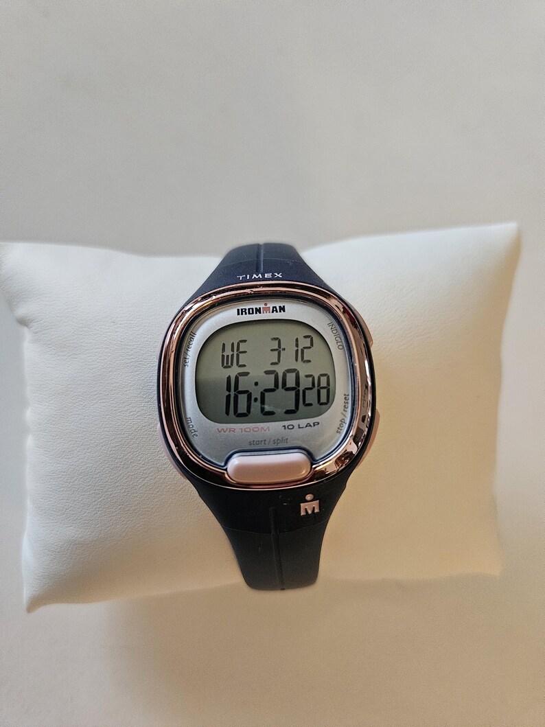 May include: A black and rose gold Timex Ironman watch with a digital display showing the time, date, and lap counter. The watch is water resistant to 300 meters.