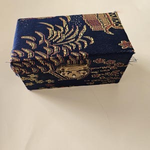 May include: A small, rectangular jewelry box with a hinged lid. The box is covered in a dark blue fabric with a gold and red floral pattern. It has a silver-colored clasp.