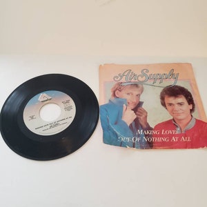 May include: A black vinyl record with the text "Making Love Out Of Nothing At All" and "Air Supply" printed on the label. The record is next to a pink and white record sleeve with the text "Air Supply" and "Making Love Out Of Nothing At All" printed on it. The sleeve features a photo of two men.