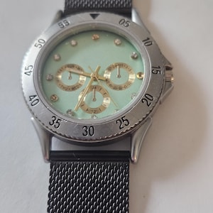 May include: A wristwatch with a mint green dial, gold-tone hands, and small crystal hour markers. The watch features three sub-dials for day, date, and 24-hour time, a silver-tone bezel with black numerals, and a black mesh metal strap.
