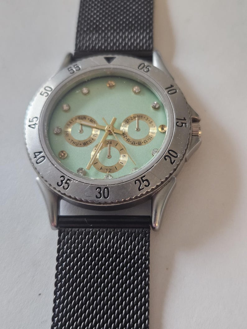 May include: A wristwatch with a mint green dial, gold-tone hands, and small crystal hour markers. The watch features three sub-dials for day, date, and 24-hour time, a silver-tone bezel with black numerals, and a black mesh metal strap.