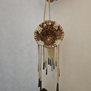 May include: A small, round, woven basket with a brown, textured surface. The basket features a light-colored leather strap for hanging and several light-colored leather fringe strips attached to the rim, each tipped with a metallic silver cone.