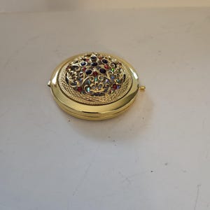 Peut inclure: Un miroir de poche rond de couleur dorée. Le dessus est orné d'un motif orné avec des pierres précieuses colorées dans des tons rouges, bleus et irisés. Le compact a une bordure texturée et une ouverture à charnière.
