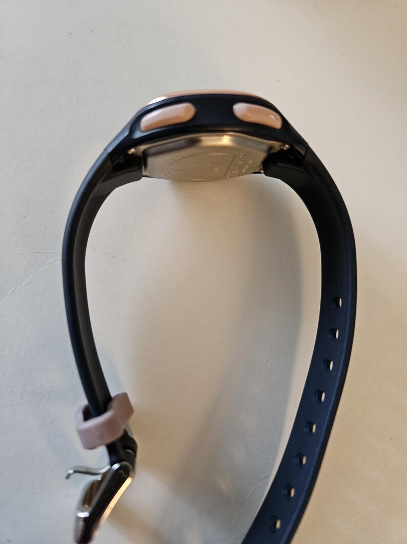 May include: A black and rose gold digital watch with a black silicone band. The watch has two buttons on the side and a silver buckle.