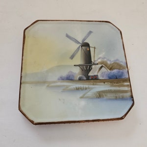 May include: A square porcelain plate with a hand-painted scene of a windmill in a snowy landscape. The windmill is black with white blades and a brown roof. The scene is painted in shades of blue, white, and brown.